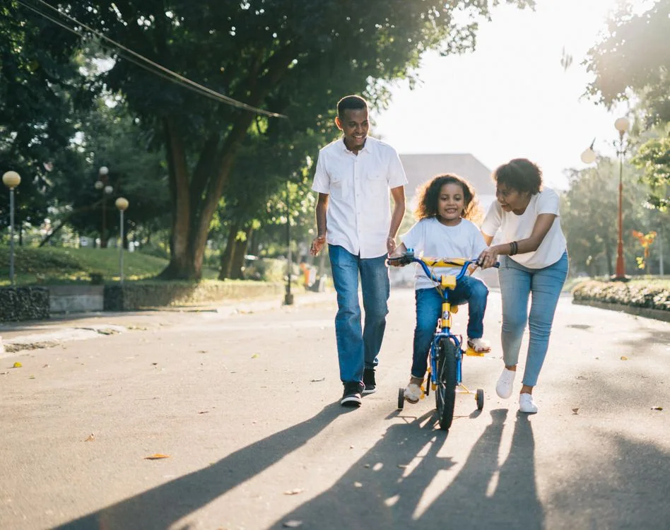 Two parents helping their child ride a bicycle along a sunlit path