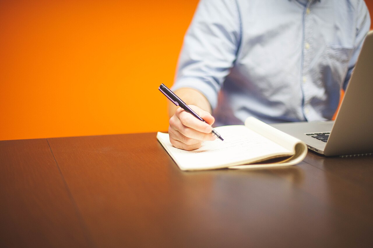 A person taking handwritten notes beside an open laptop.